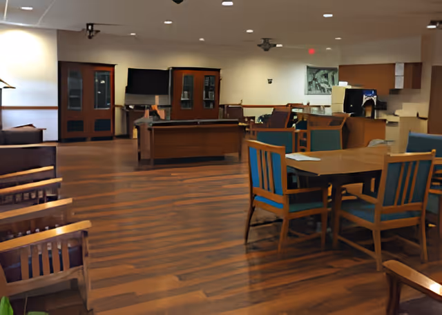 Spacious senior living common area with wooden flooring, multiple wooden chairs with blue cushions, wooden tables, and cabinets along the walls. The room is well-lit with ceiling lights and has a cozy, inviting atmosphere.