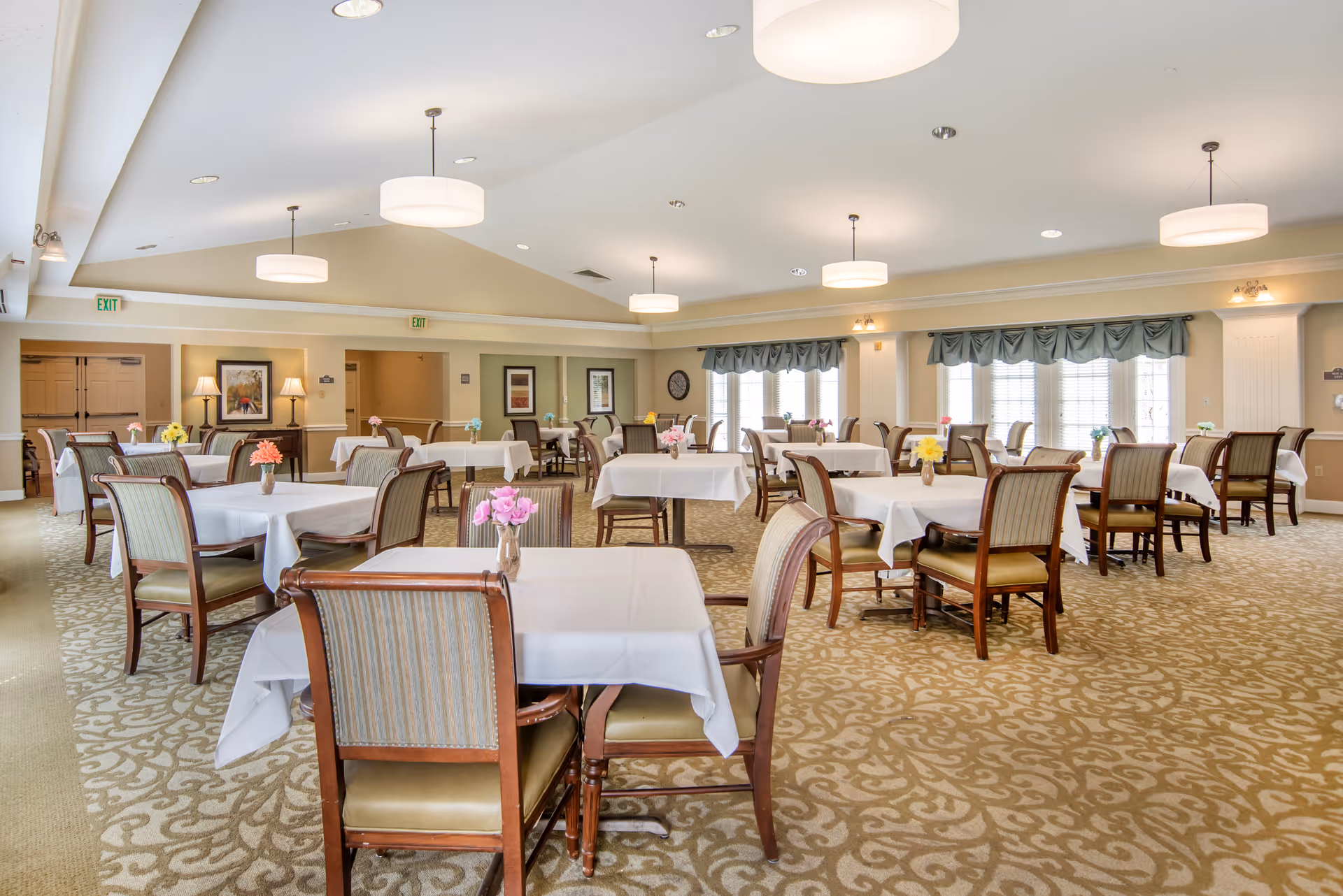 A spacious dining room with multiple tables covered in white tablecloths, each adorned with small vases of colorful flowers. The room features patterned carpet, cushioned wooden chairs, large windows with valances, and ceiling lights providing bright illumination.