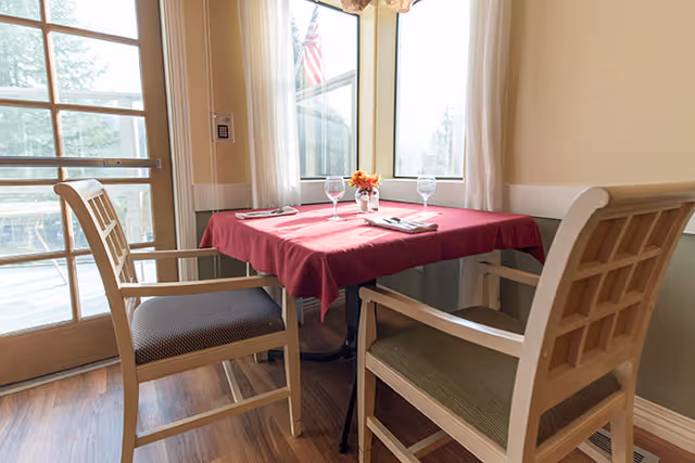 A small dining table covered with a red tablecloth set for two with two chairs, two water glasses, napkins, and a small flower arrangement. The table is positioned near large windows and a glass door letting in natural light.