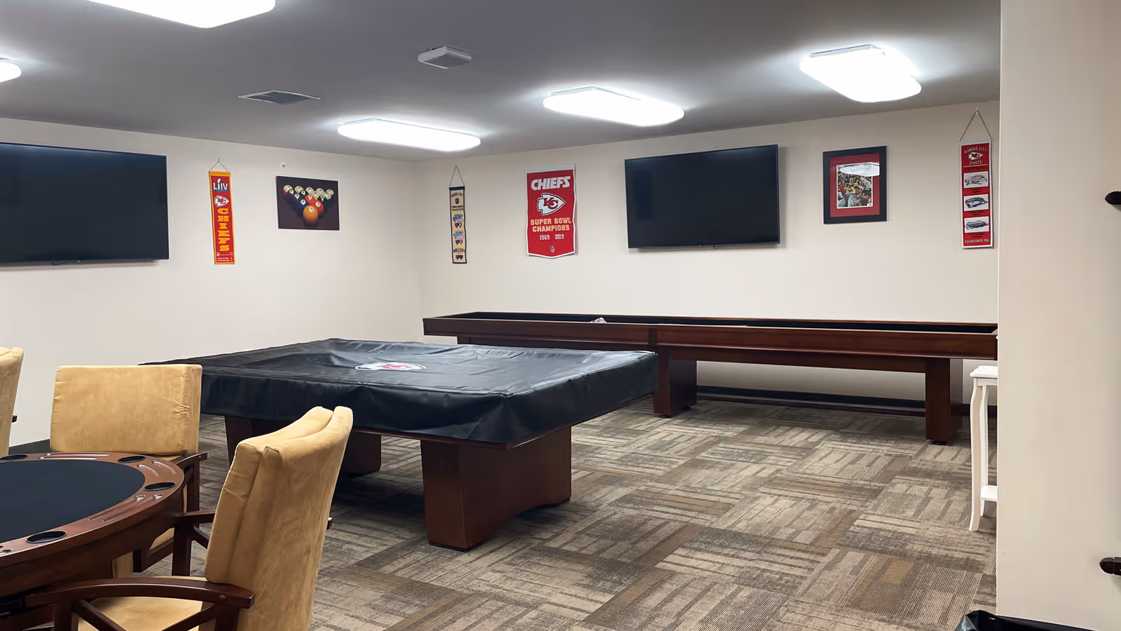 Interior view of a recreational room featuring a covered pool table, a shuffleboard table, a round poker table with four beige chairs, two wall-mounted flat-screen TVs, and sports-themed wall decorations including Kansas City Chiefs banners and pictures.