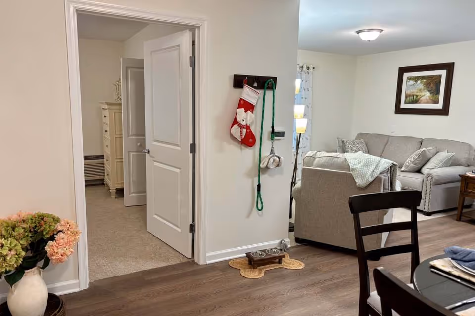 Interior view of a senior living facility showing a cozy living room with a gray sofa, armchair, and a framed picture on the wall. A dining table with chairs is partially visible in the foreground. There is an open doorway leading to a bedroom with a dresser. A Christmas stocking and dog leash hang on a wall near a pet feeding station on the floor.
