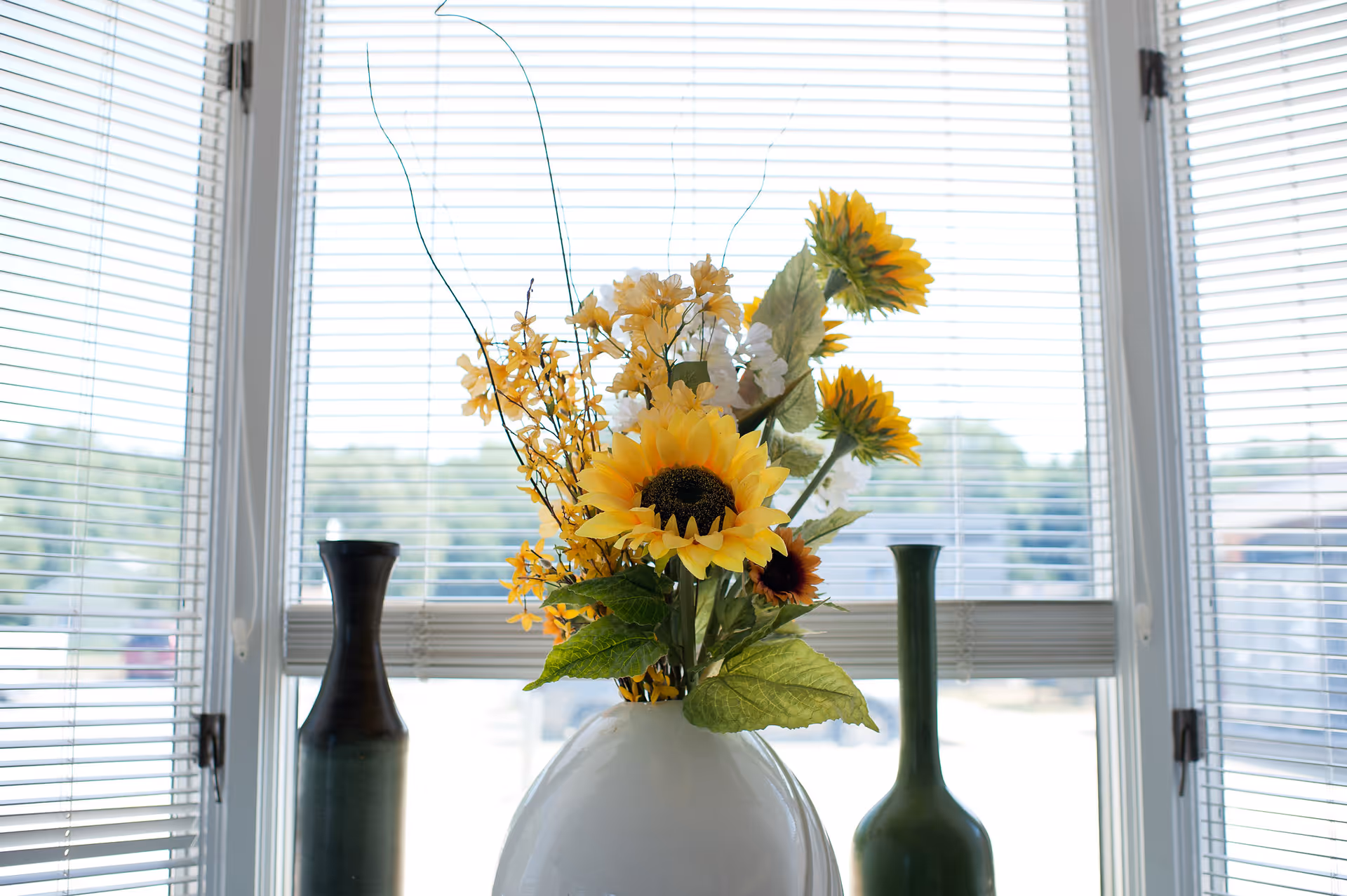A vase of sunflowers and yellow flowers on a windowsill with blinds and two tall green vases.