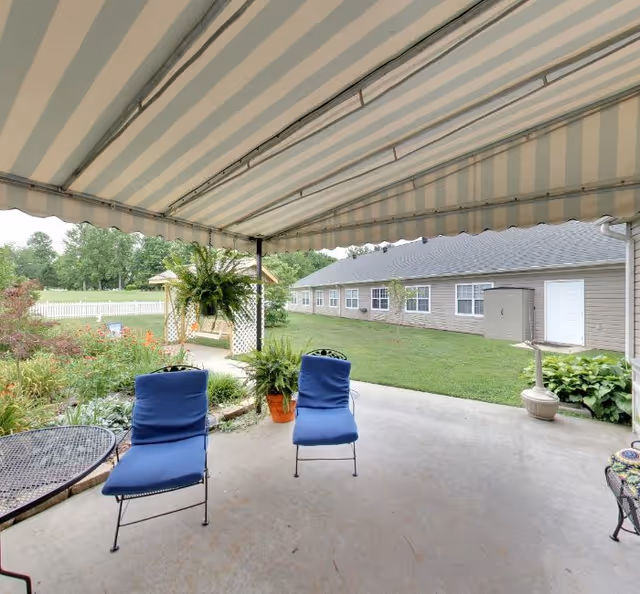 Covered patio with a striped awning, two blue cushioned chairs, potted plants and a lawn with a building in the background.