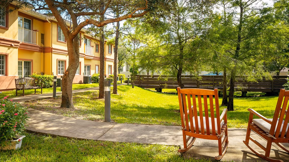 Outdoor garden area at Wickshire Tamarac featuring a paved walkway, green grass, several trees, wooden benches, and two wooden rocking chairs in the foreground. The building with yellow and brown exterior walls and windows is visible on the left side.