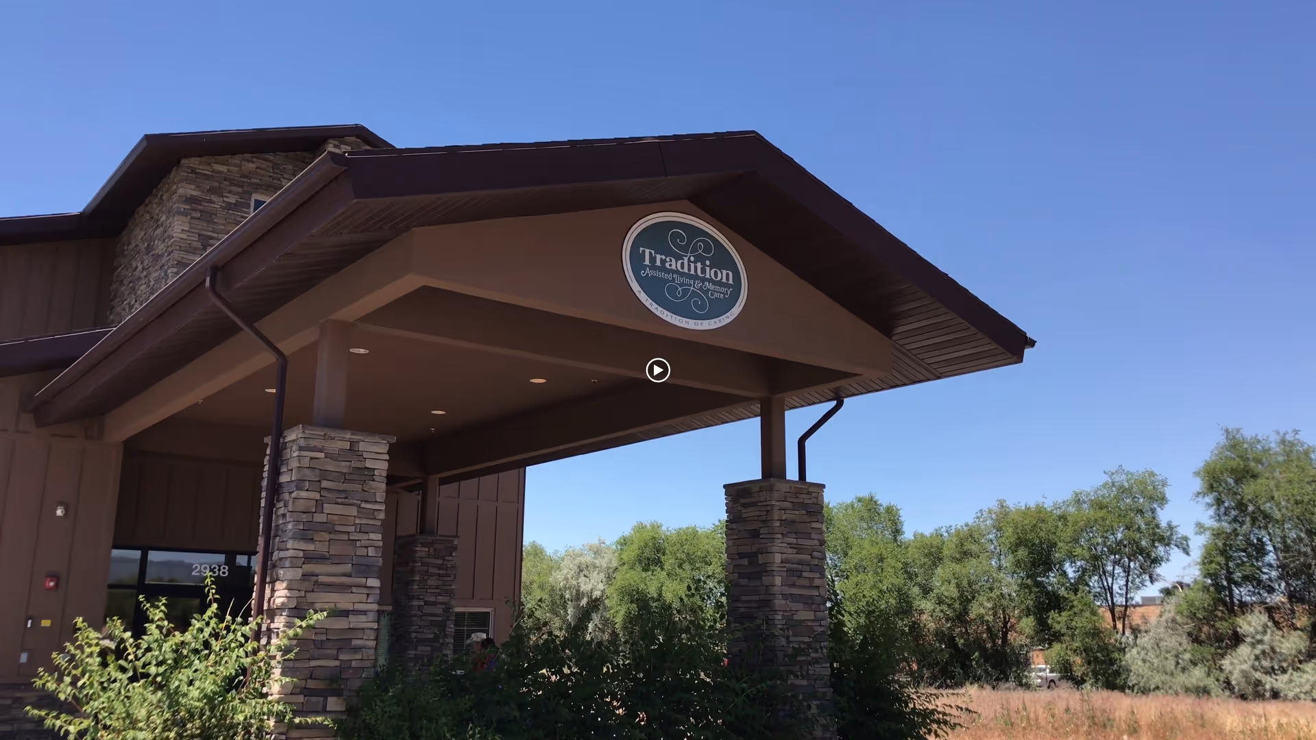 Front exterior view of Tradition Assisted Living facility with a covered entrance supported by stone pillars, surrounded by greenery and trees under a clear blue sky.