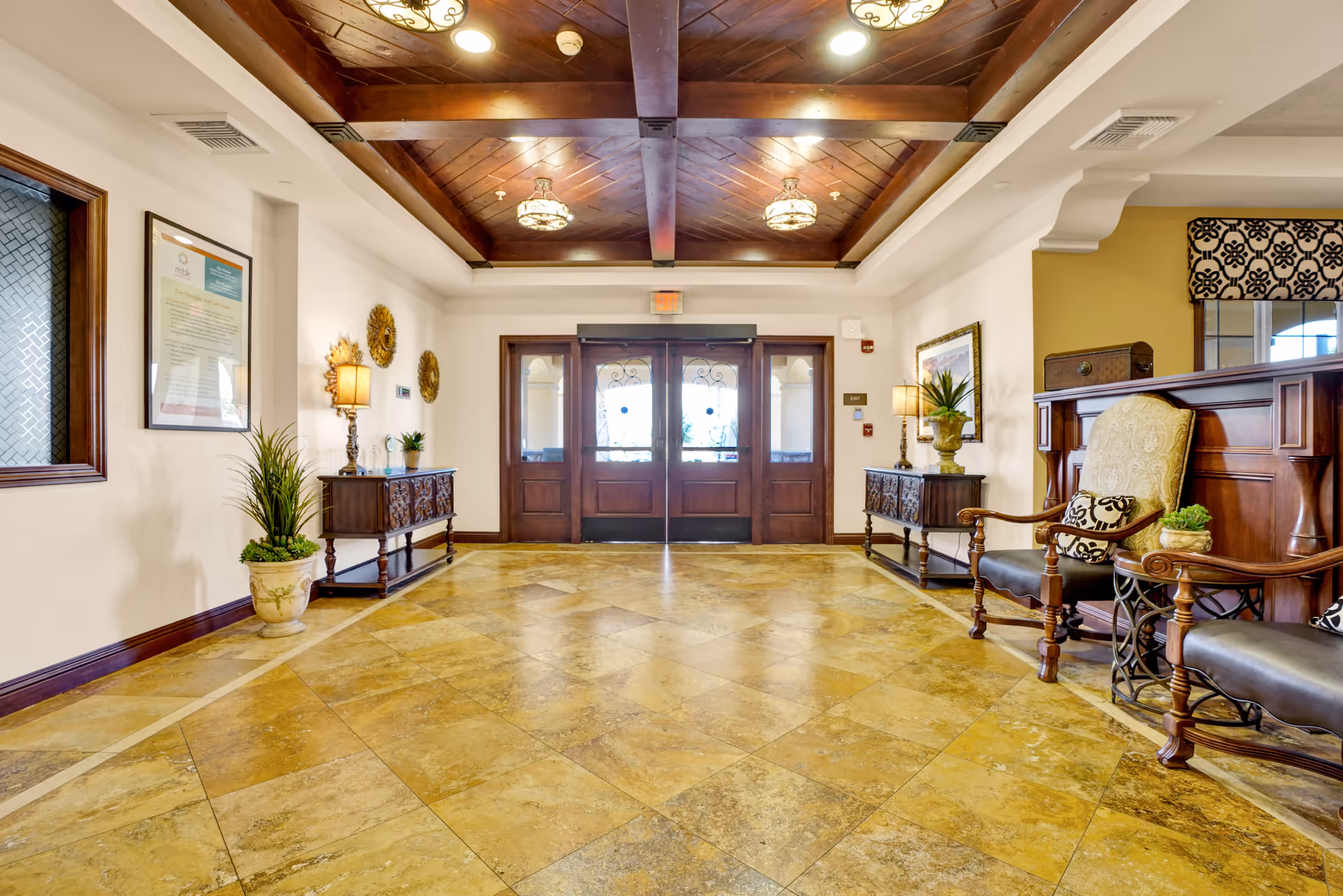 A spacious and elegant lobby area with a tiled floor, wooden ceiling beams, and decorative light fixtures. There are two wooden side tables with lamps and plants on either side of the double glass entrance doors. On the right, there are two cushioned wooden chairs with patterned pillows next to a wooden reception desk. The walls are decorated with framed artwork and ornamental wall hangings.