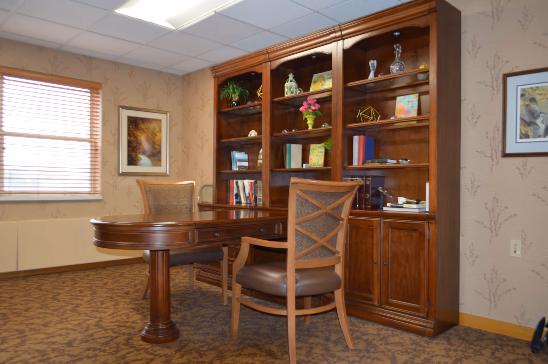 A cozy office or study room with a large wooden desk and two matching chairs. Behind the desk is a tall wooden bookshelf filled with books, decorative items, and plants. The room has patterned carpet, wallpaper with subtle designs, a window with wooden blinds, and framed artwork on the walls.