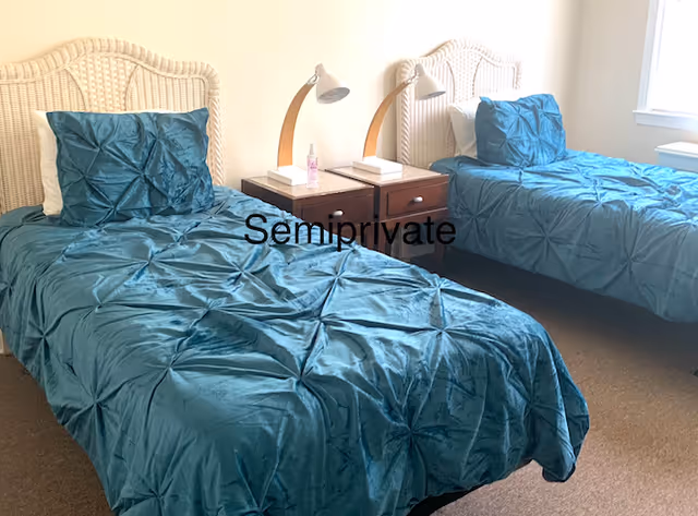 A bedroom with two twin beds featuring white wicker headboards and teal comforters. Between the beds is a dark wooden nightstand with two white lamps and a small bottle. The room is well-lit with natural light coming from a window on the right.