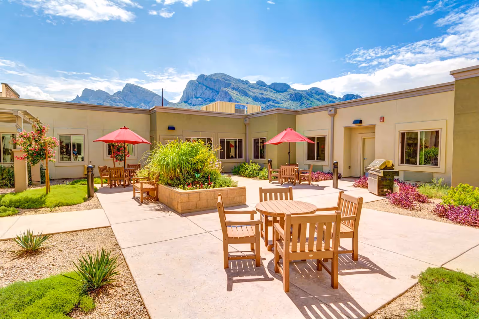 Outdoor courtyard area at Catalina Springs Memory Care with wooden tables and chairs, red umbrellas, a built-in planter with greenery, and a barbecue grill. The courtyard is surrounded by a single-story building with windows and doors, and mountains are visible in the background under a partly cloudy blue sky.