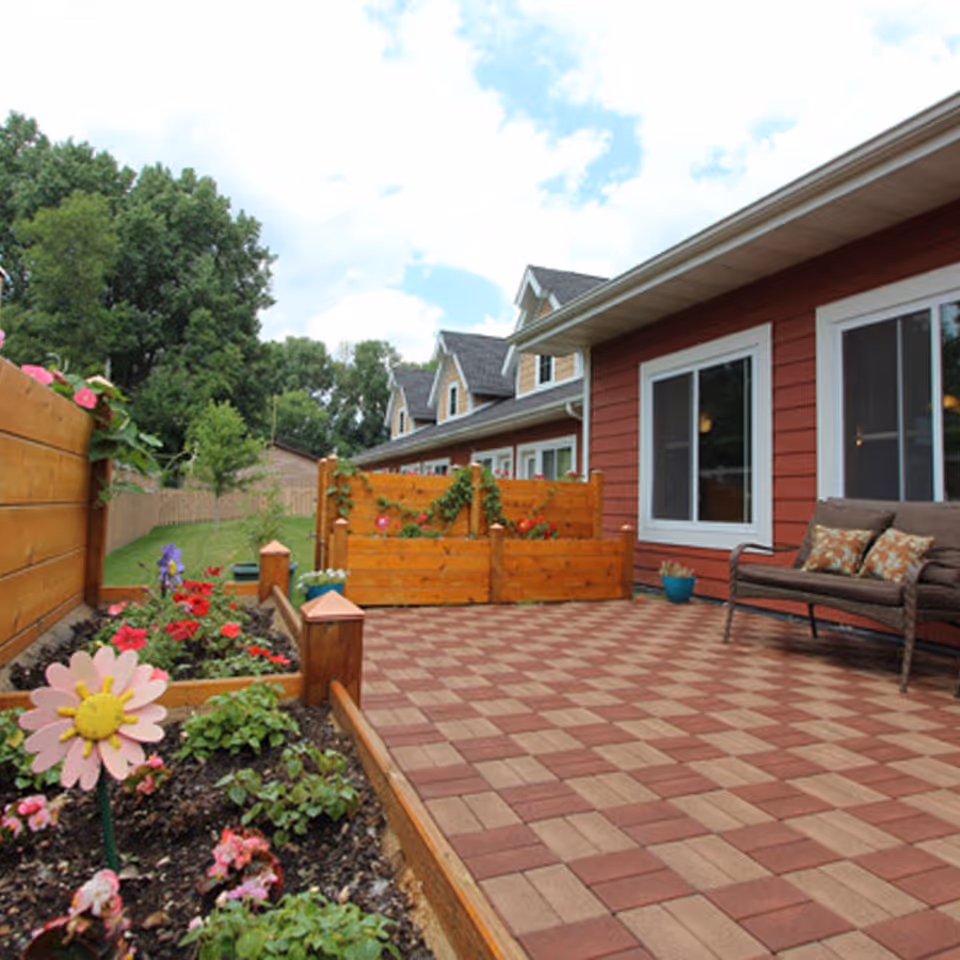 Paved patio with wooden planters, flowers and a bench beside a red-sided assisted living building and landscaped yard.