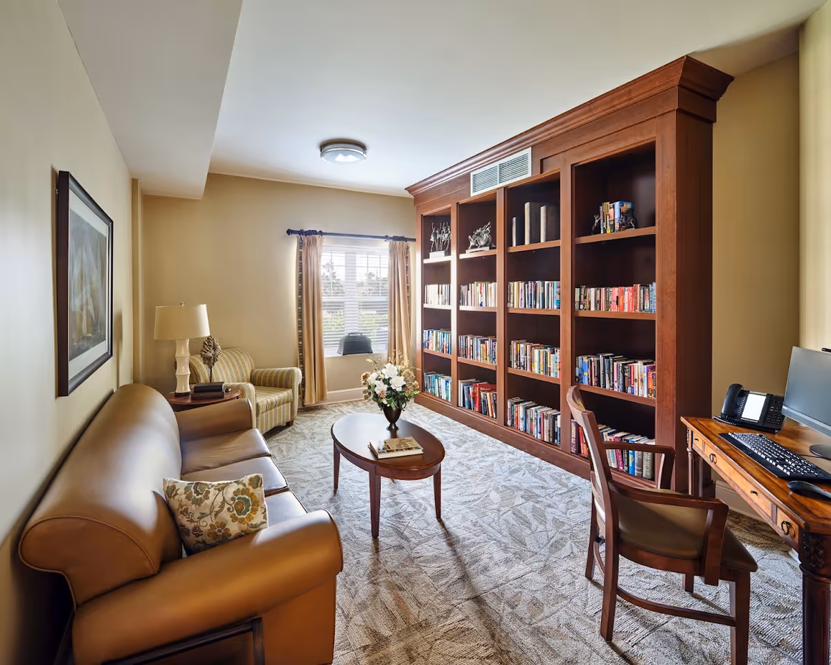 Comfortable senior living common room with a leather sofa, armchair, coffee table, large built-in bookshelves, and a desk with a computer.