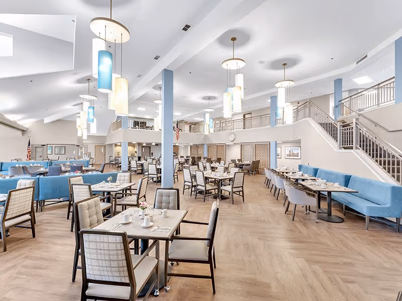 Spacious dining room in a senior living facility with multiple tables and chairs arranged neatly. The room features light wood flooring, blue cushioned bench seating along the walls, and modern pendant lights hanging from a high ceiling. A staircase and balcony area are visible in the background, along with American flags and framed pictures on the walls.
