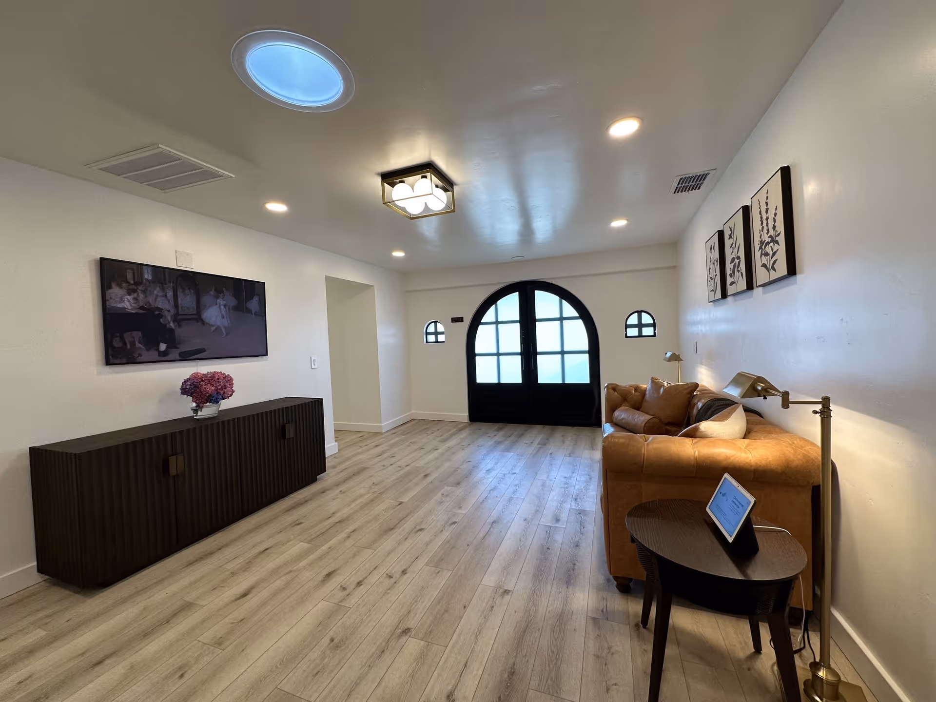 A modern interior room with light wood flooring, white walls, and a round skylight on the ceiling. There is a dark wooden cabinet with a vase of flowers on top against the left wall, and a large flat-screen TV mounted above it. On the right side, there is a brown leather sofa with pillows, a small round side table holding a digital device, and a floor lamp. The far wall features a large black double door with frosted glass panels and two small arched windows on either side. Three framed botanical prints hang on the right wall above the sofa.