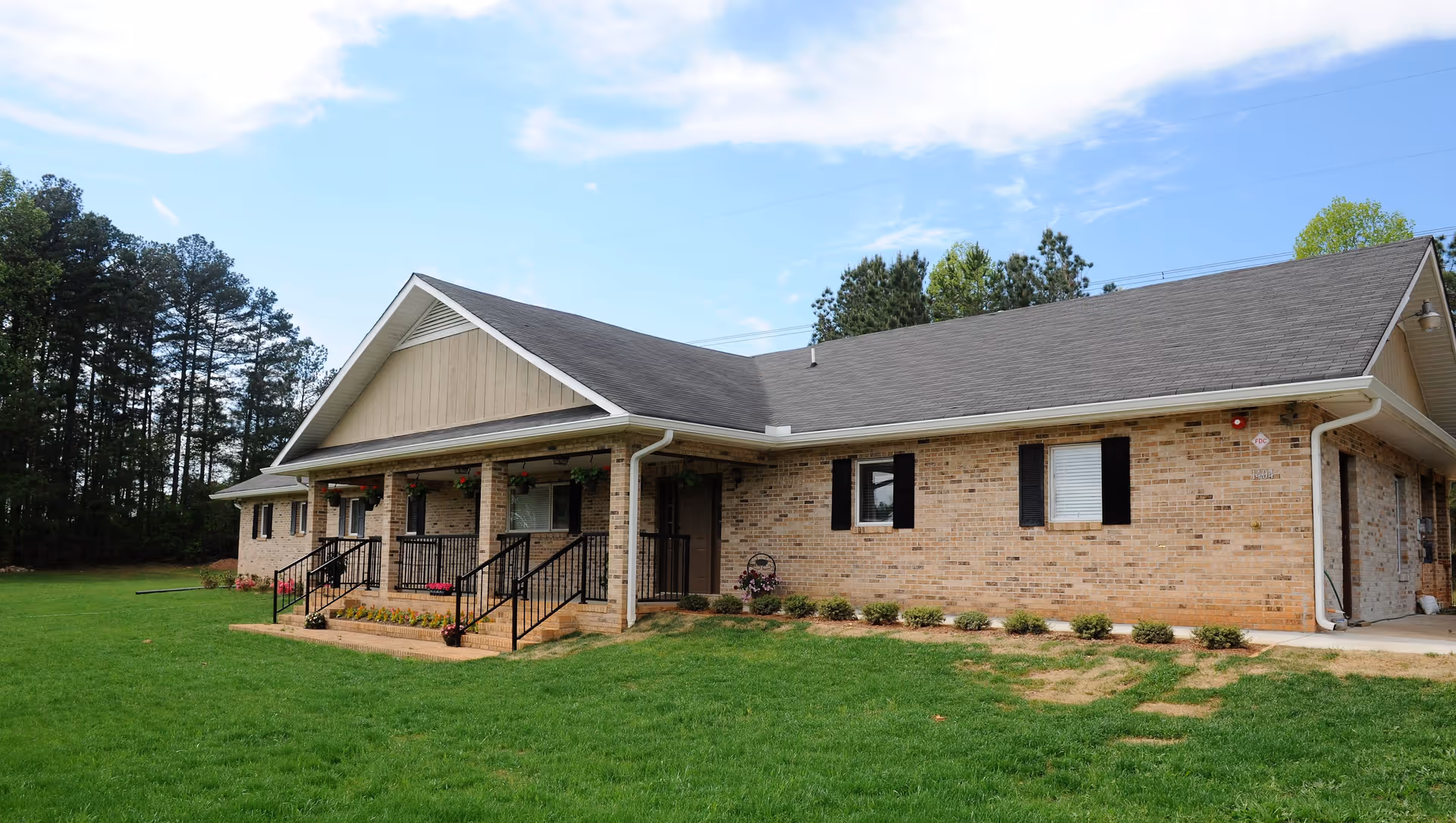Single-story brick building with a gabled roof, a covered porch with black railings, and a well-maintained green lawn in front. The building is surrounded by trees under a partly cloudy sky.