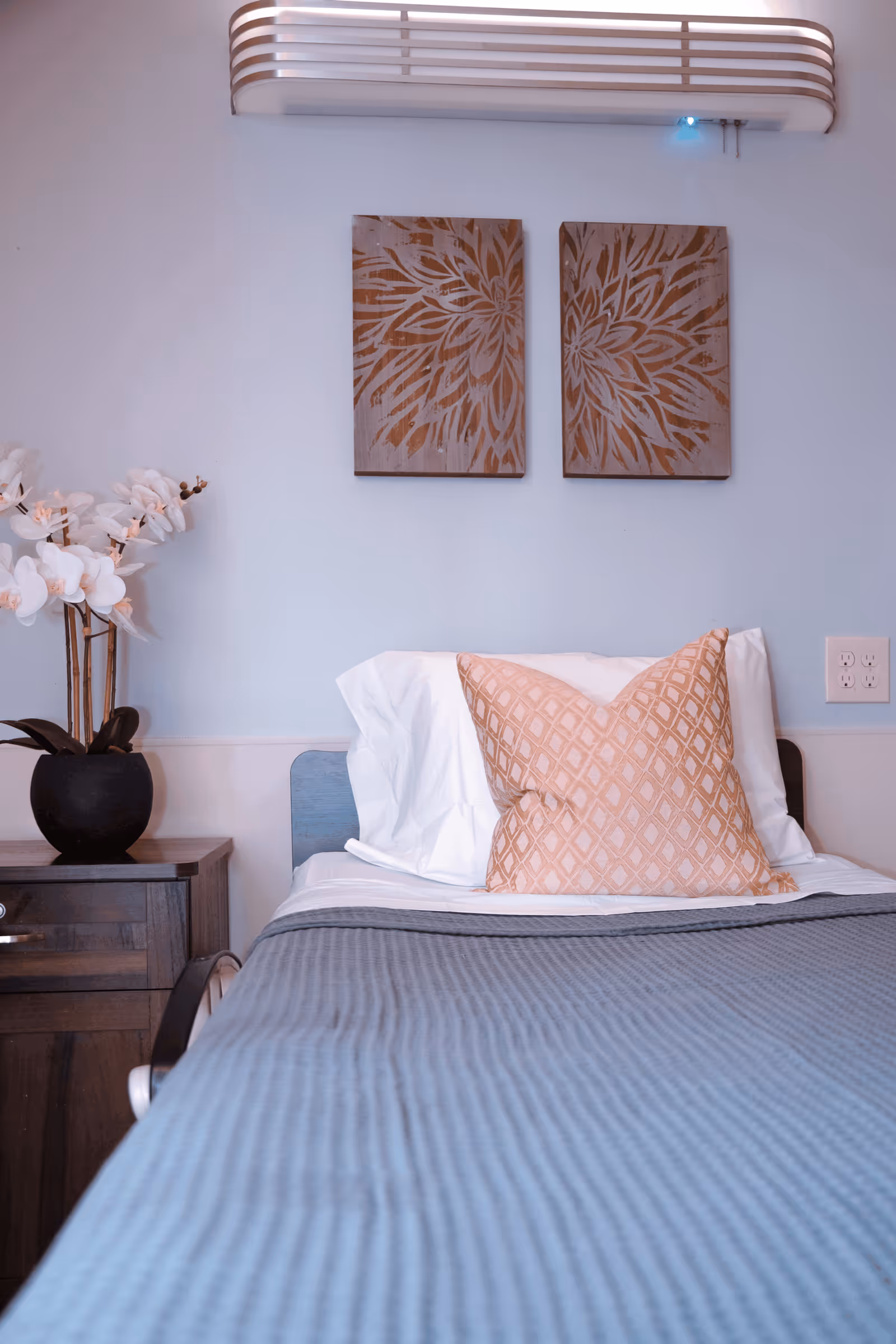 A neatly made bedroom with a bed, white linens, a peach patterned pillow, a wooden nightstand with a plant, and two-panel wall art above the headboard.
