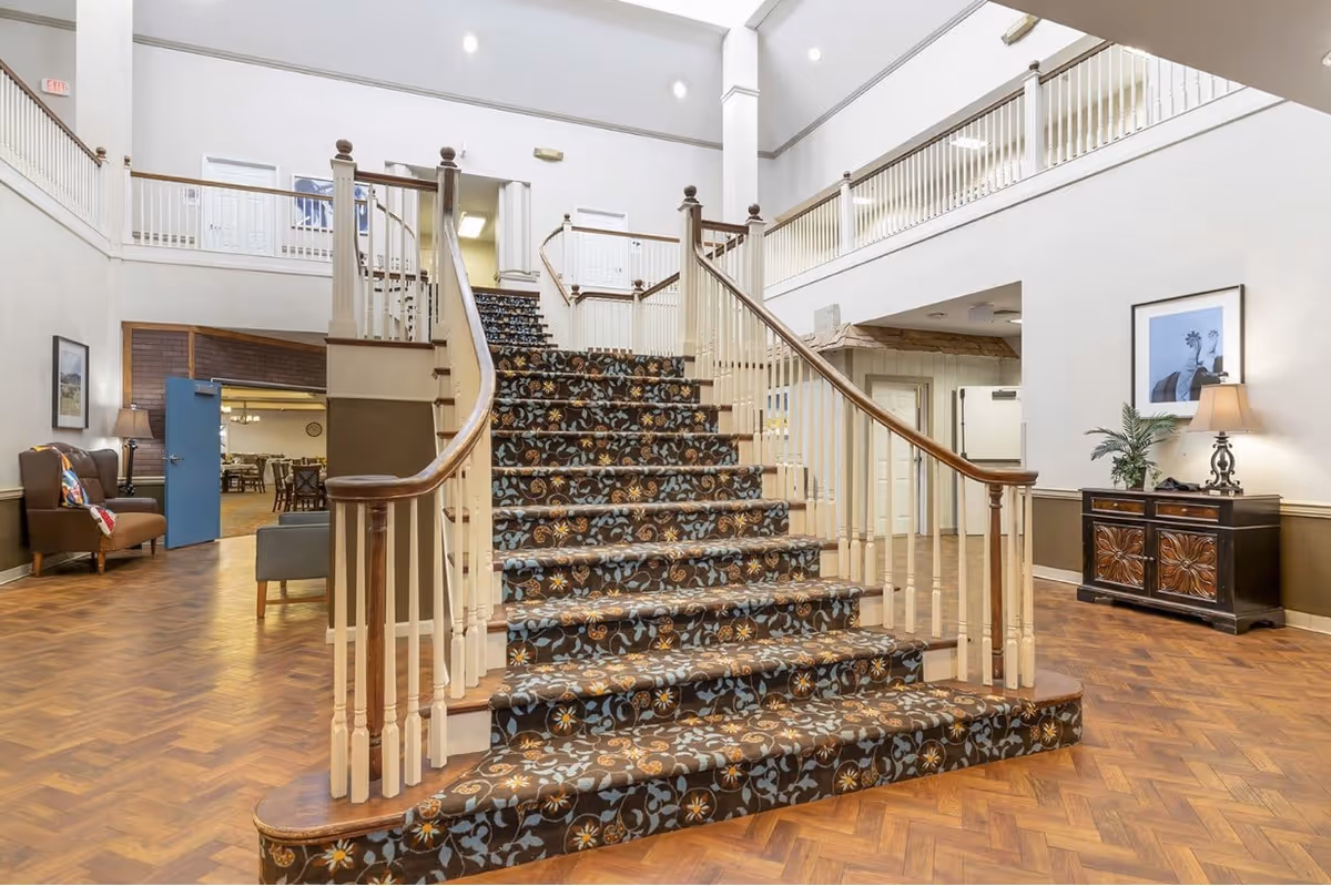 Interior view of a senior living facility featuring a wide staircase with patterned carpet and wooden handrails. The area has a high ceiling with white walls and a wooden floor. To the left, there is a seating area with armchairs and a lamp, and in the background, a dining area with tables and chairs is visible. On the right side, there is a decorative cabinet with a lamp and a framed picture above it.