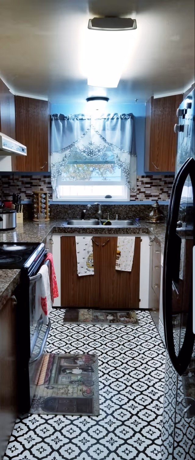 A compact kitchen with wooden cabinets, granite countertops, and a patterned black and white tiled floor. There is a double sink beneath a window with decorative curtains. Kitchen towels hang on the cabinet doors under the sink. Appliances include a black stove with a red towel hanging on the handle and a black refrigerator. The backsplash features a mosaic tile design in shades of brown and beige.
