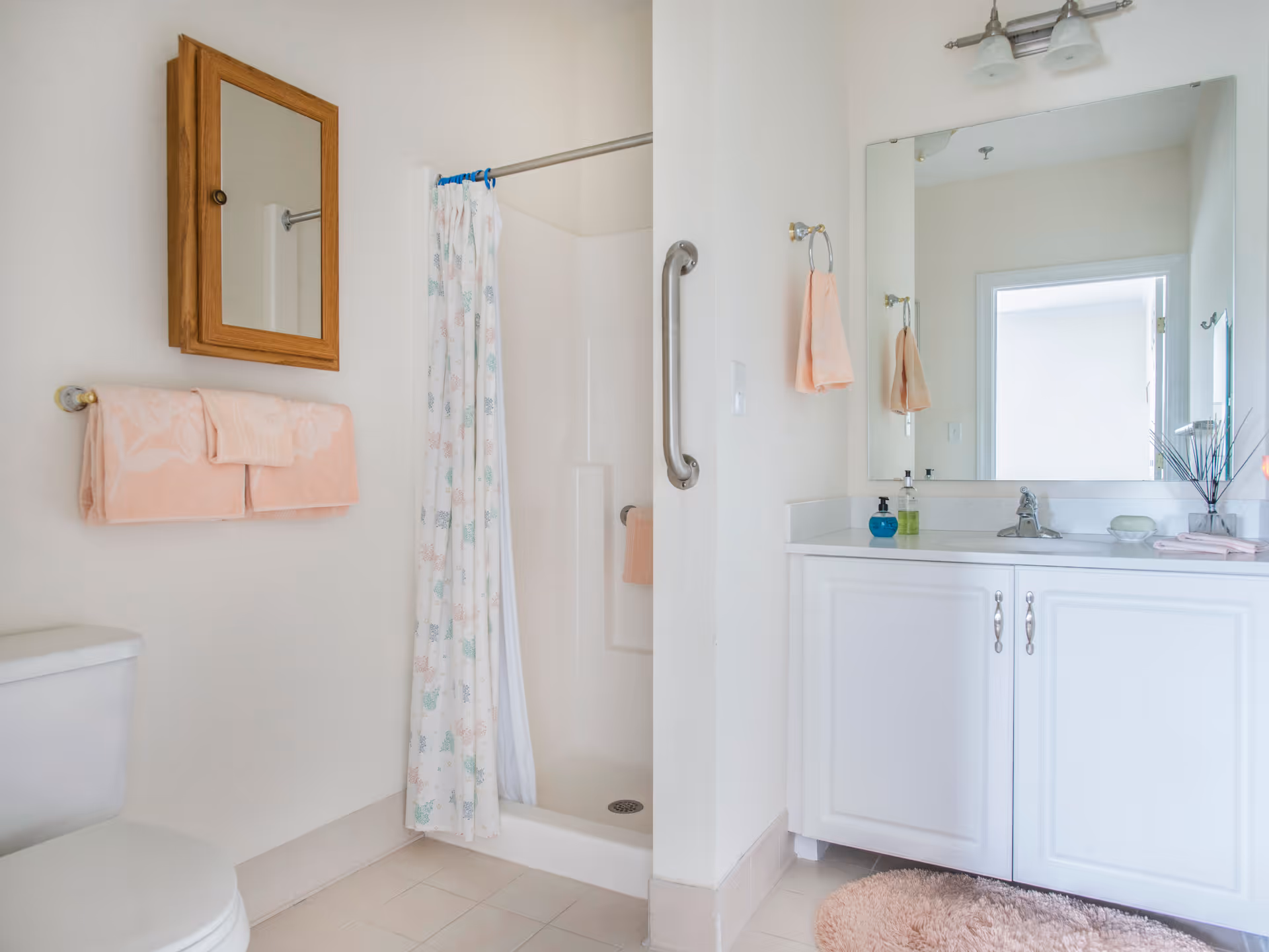 A clean bathroom with a white toilet, a shower with a floral shower curtain, a wooden framed mirror above a towel rack holding pink towels, and a white vanity with a large mirror, sink, and various toiletries. A pink bath mat is on the floor in front of the vanity.