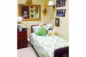 Small decorated bedroom with a single bed in green floral bedding, stuffed animals on the bed, a dresser with a lamp, a wall mirror and framed photos.