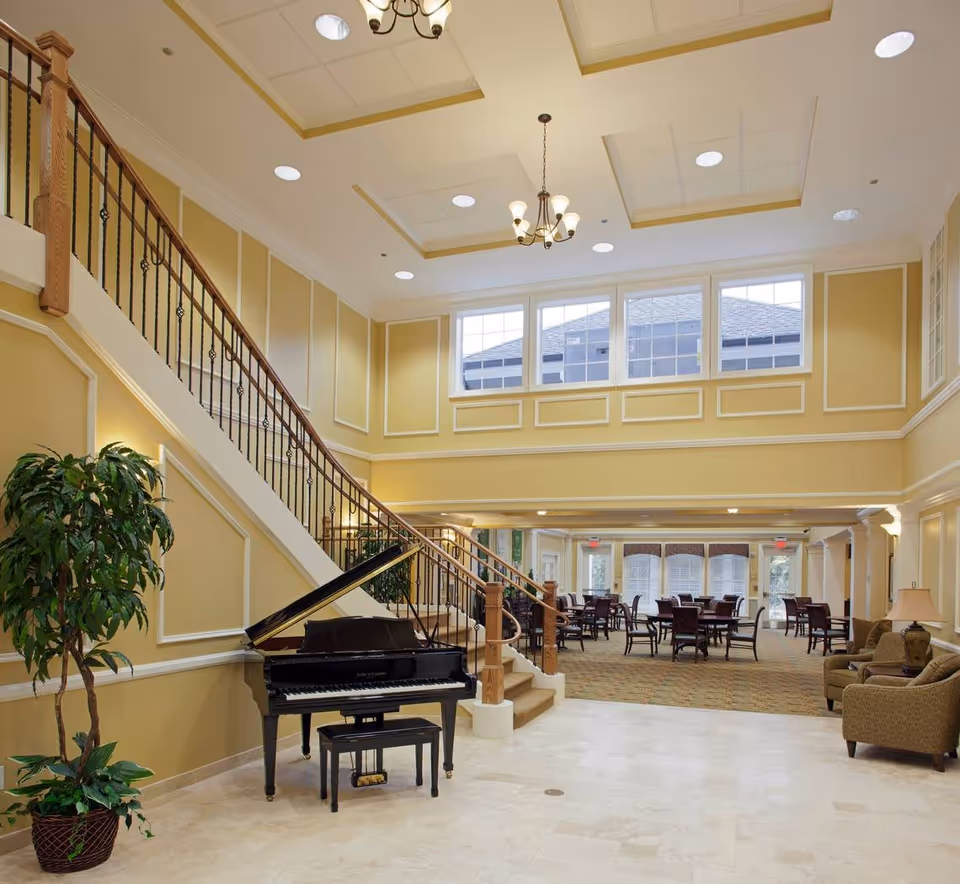 Spacious and elegant senior living facility common area with a grand piano near a staircase, large windows letting in natural light, chandeliers hanging from a high ceiling, and several tables and chairs arranged in the background for socializing or dining.