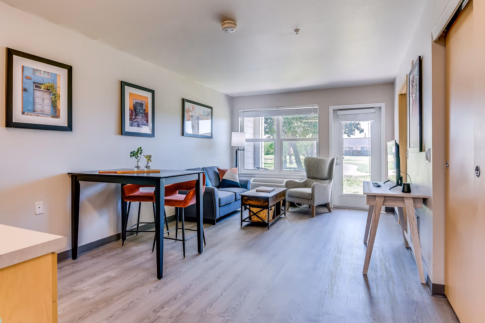 A bright and modern living room in a senior living facility featuring a gray sofa with colorful pillows, a gray armchair, a wooden coffee table with a basket underneath, a tall floor lamp, and a small dining table with two red chairs. The room has large windows and a glass door that provide a view of the outdoor greenery. Three framed pictures hang on the wall above the dining table.
