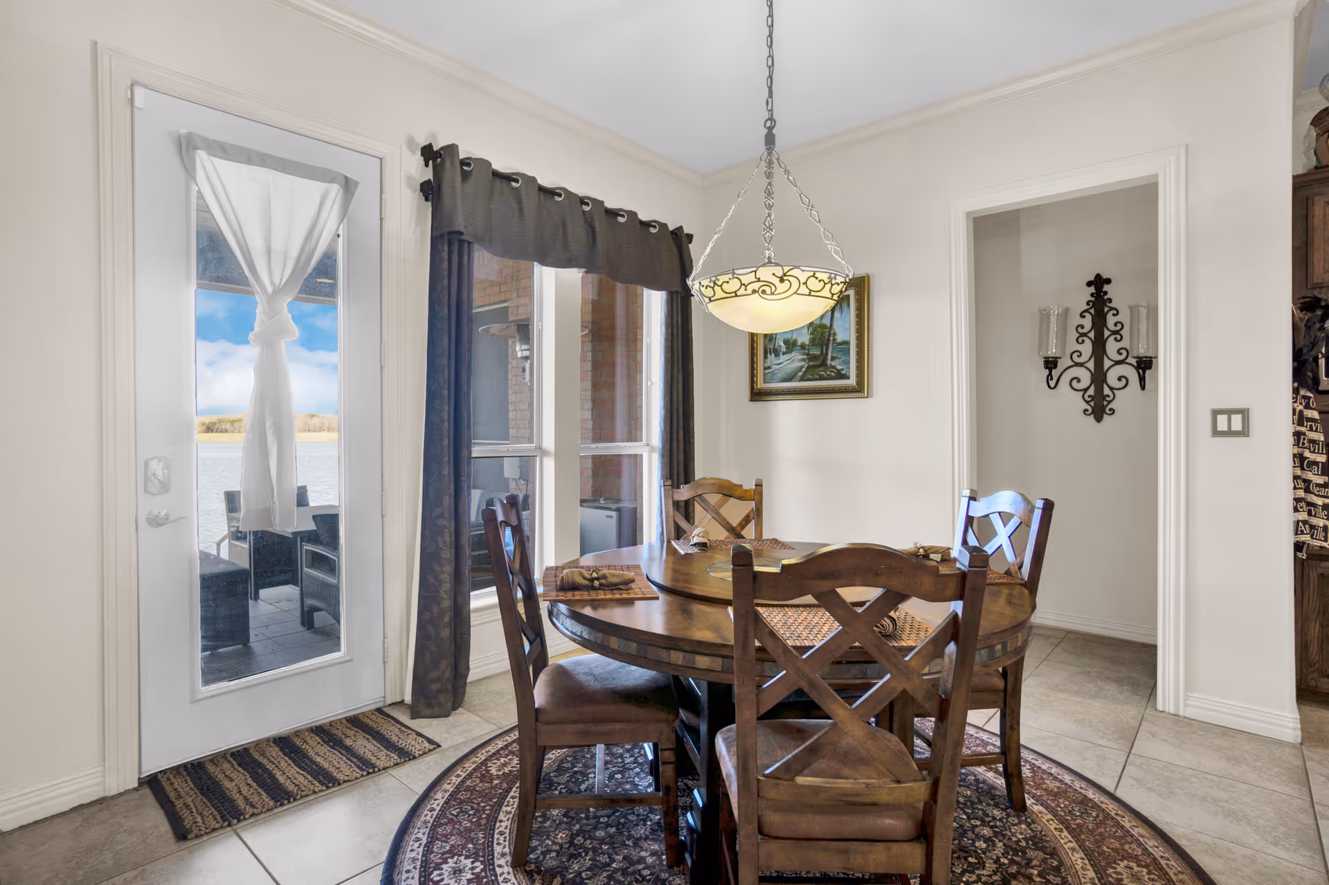 A dining area with a round wooden table and four matching chairs on a patterned circular rug. The table is set with placemats and napkins. A decorative hanging light fixture is above the table. There is a door with a white curtain tied in the center leading outside, and a window with dark curtains next to it. A framed painting and a wall-mounted candle holder are visible on the walls.
