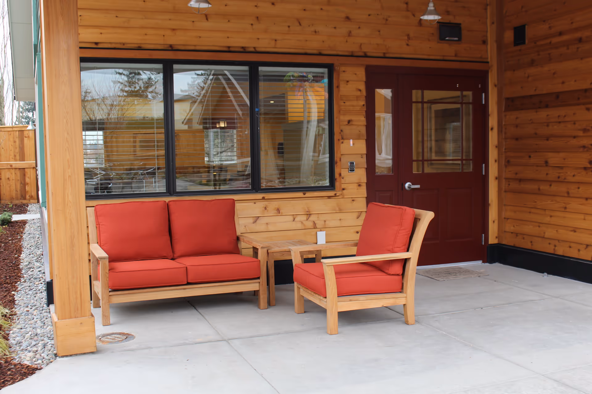 Outdoor seating area with wooden furniture including a loveseat and a chair with red cushions, a small wooden table between them, set against a wooden wall with a window and a maroon door.