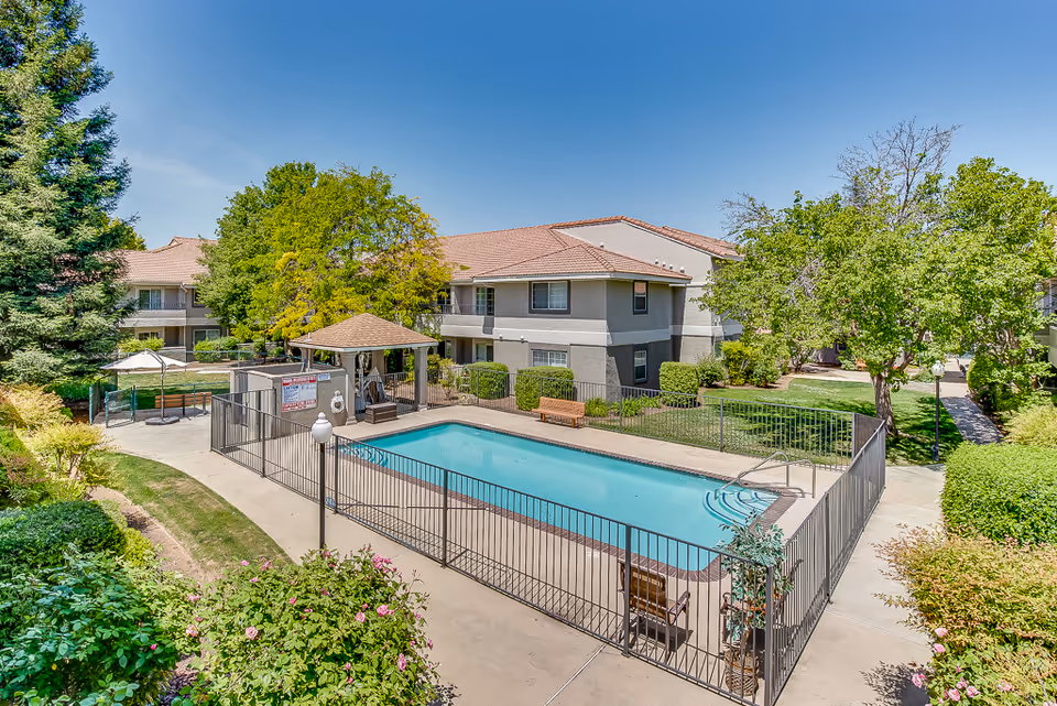 Outdoor swimming pool area surrounded by a black metal fence with a small covered seating area nearby. The pool is located in the courtyard of a two-story senior living facility building with beige walls and red-tiled roofs. There are trees, bushes, and well-maintained landscaping around the pool and building under a clear blue sky.