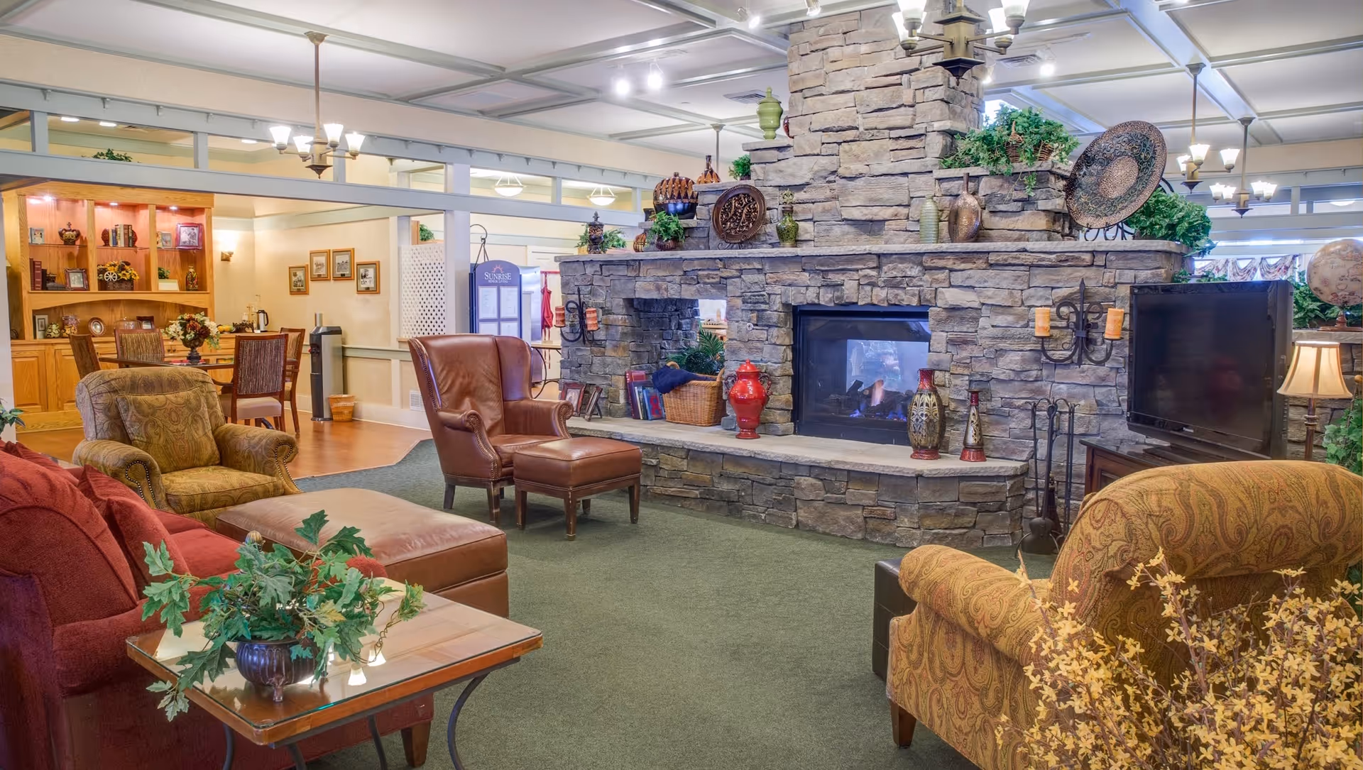 A cozy living room area in a senior living facility featuring a large stone fireplace with decorative items on the mantel. The room has various upholstered chairs and sofas arranged around the fireplace, a TV on a stand to the right, and a wooden table with a plant in the foreground. The background shows a dining area with wooden chairs and a cabinet with shelves displaying decorative items.