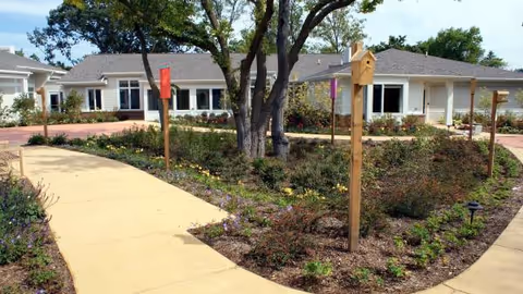 Curved concrete walkway winds through landscaped beds toward a single-story senior living facility.