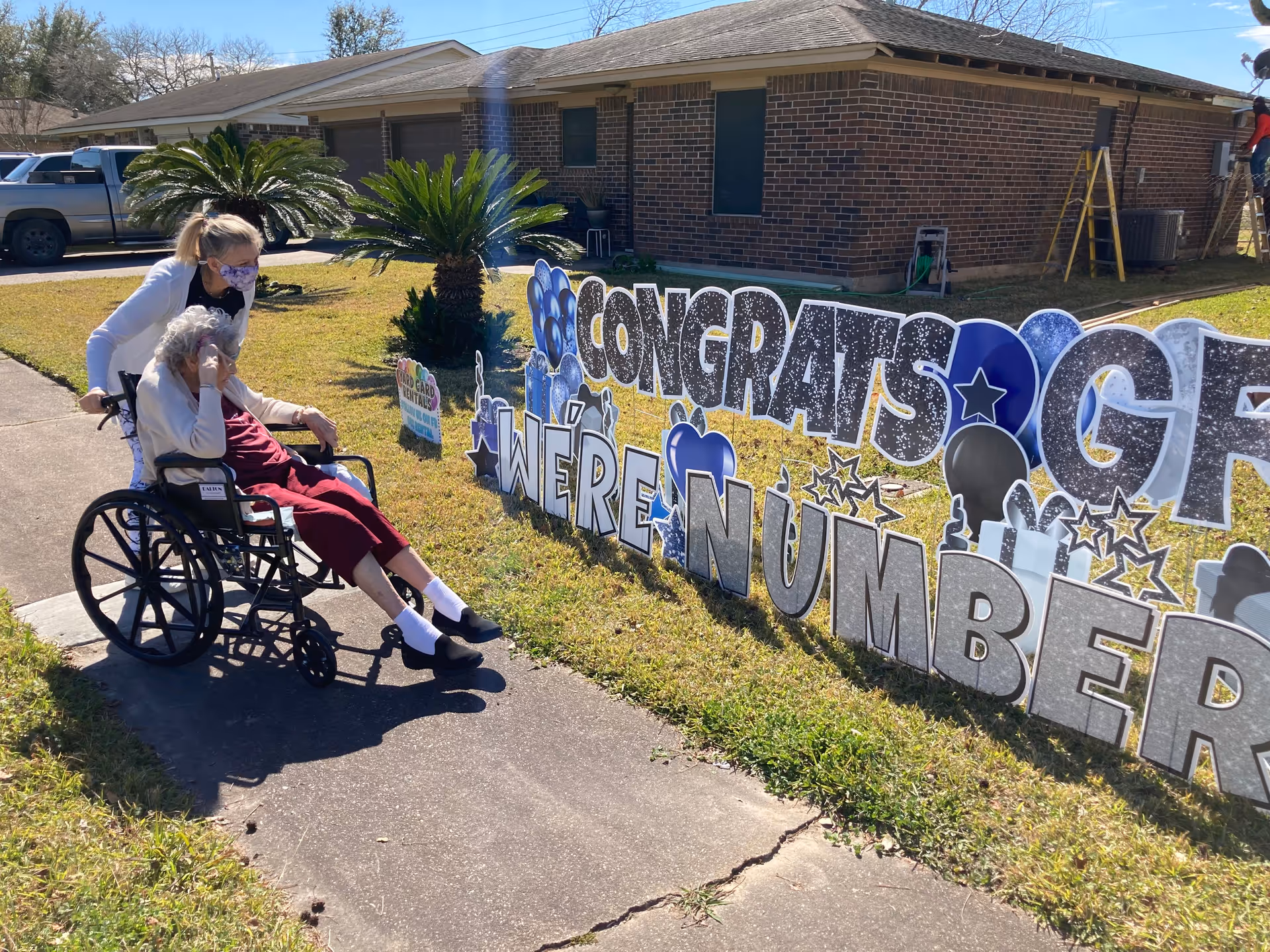 An elderly woman in a wheelchair is being pushed by a caregiver wearing a face mask along a sidewalk next to a lawn. On the lawn, there is a large congratulatory sign that reads 'CONGRATS WE'RE NUMBER' with decorative stars and balloons. The background shows a brick building and some parked vehicles.