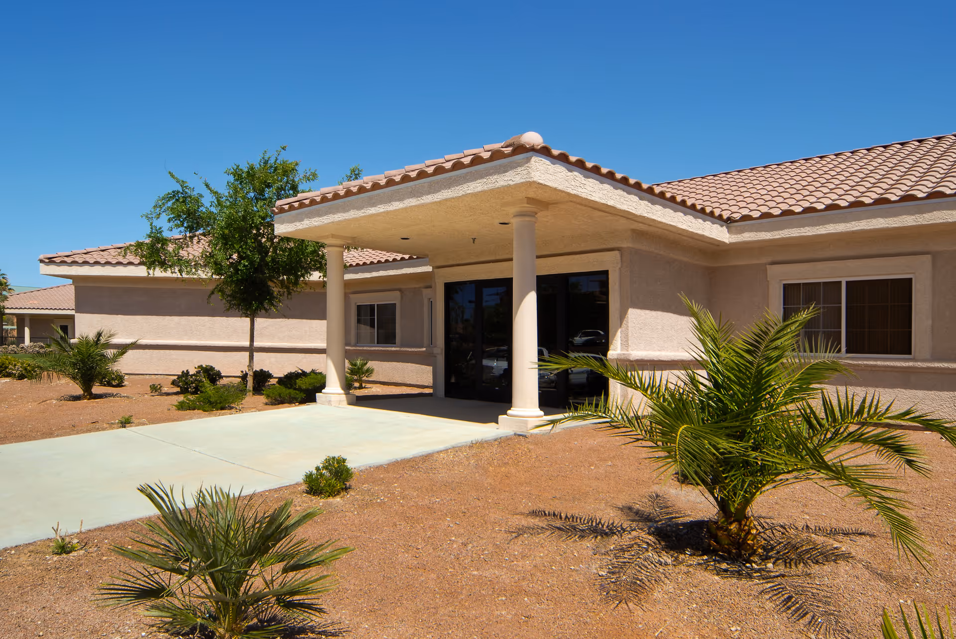 Exterior view of a single-story building with a covered entrance supported by two columns, surrounded by desert landscaping with small palm trees and shrubs under a clear blue sky.