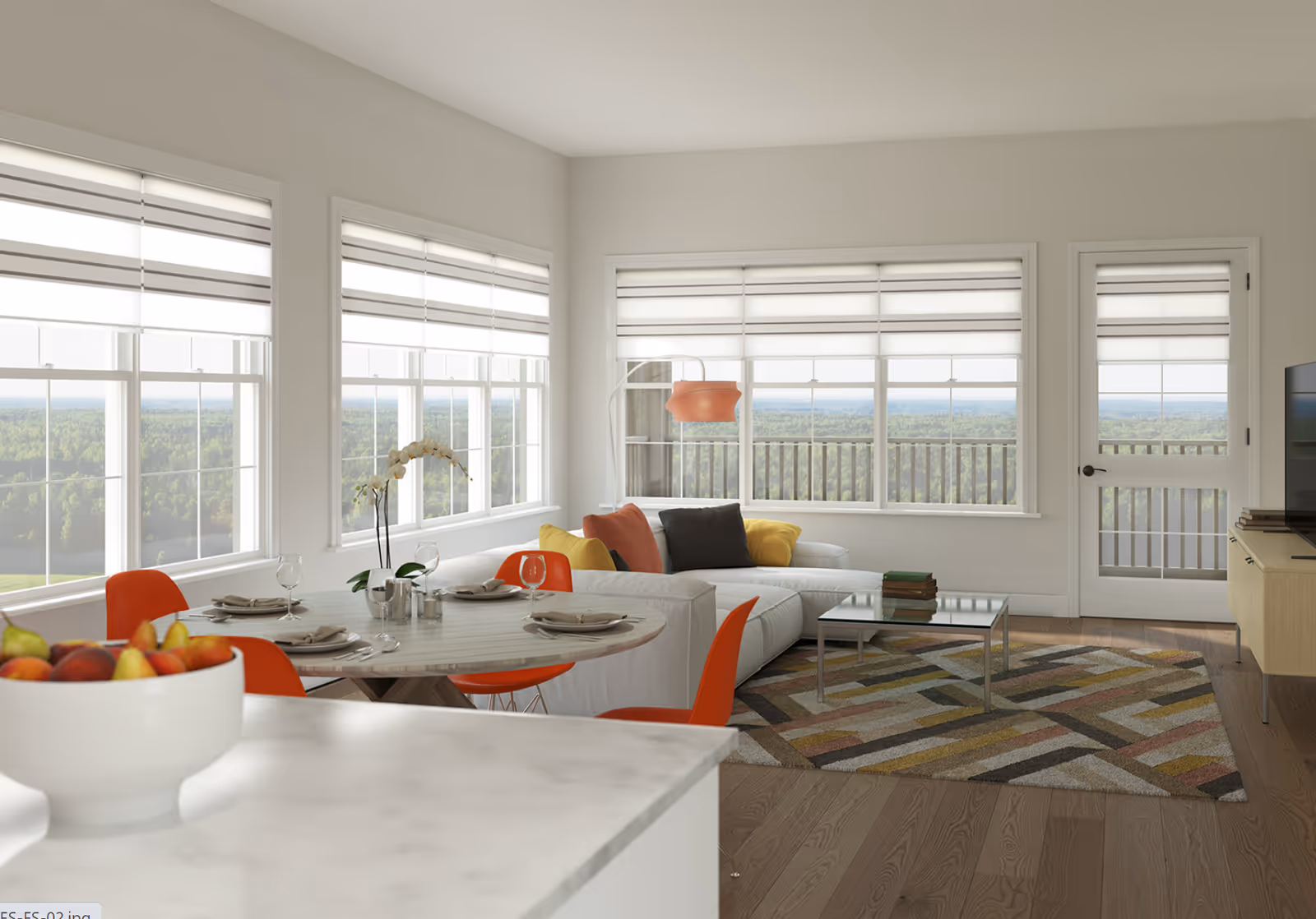 Bright and modern living room and dining area with large windows showing a scenic forest view. The room features a white sectional sofa with colorful cushions, a patterned rug, a glass coffee table, a round dining table set with plates and glasses, and orange chairs. A bowl of fruit is on a marble countertop in the foreground.