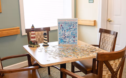 A table with an unfinished jigsaw puzzle, two lighthouse figurines and a puzzle box surrounded by four chairs near a window.