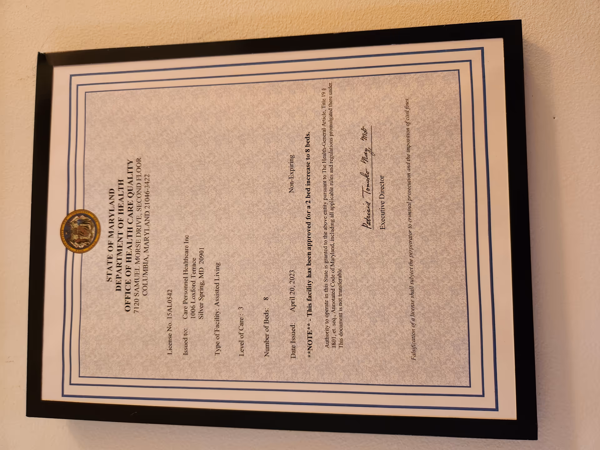Framed certificate with blue borders and an official seal hanging on a beige wall.