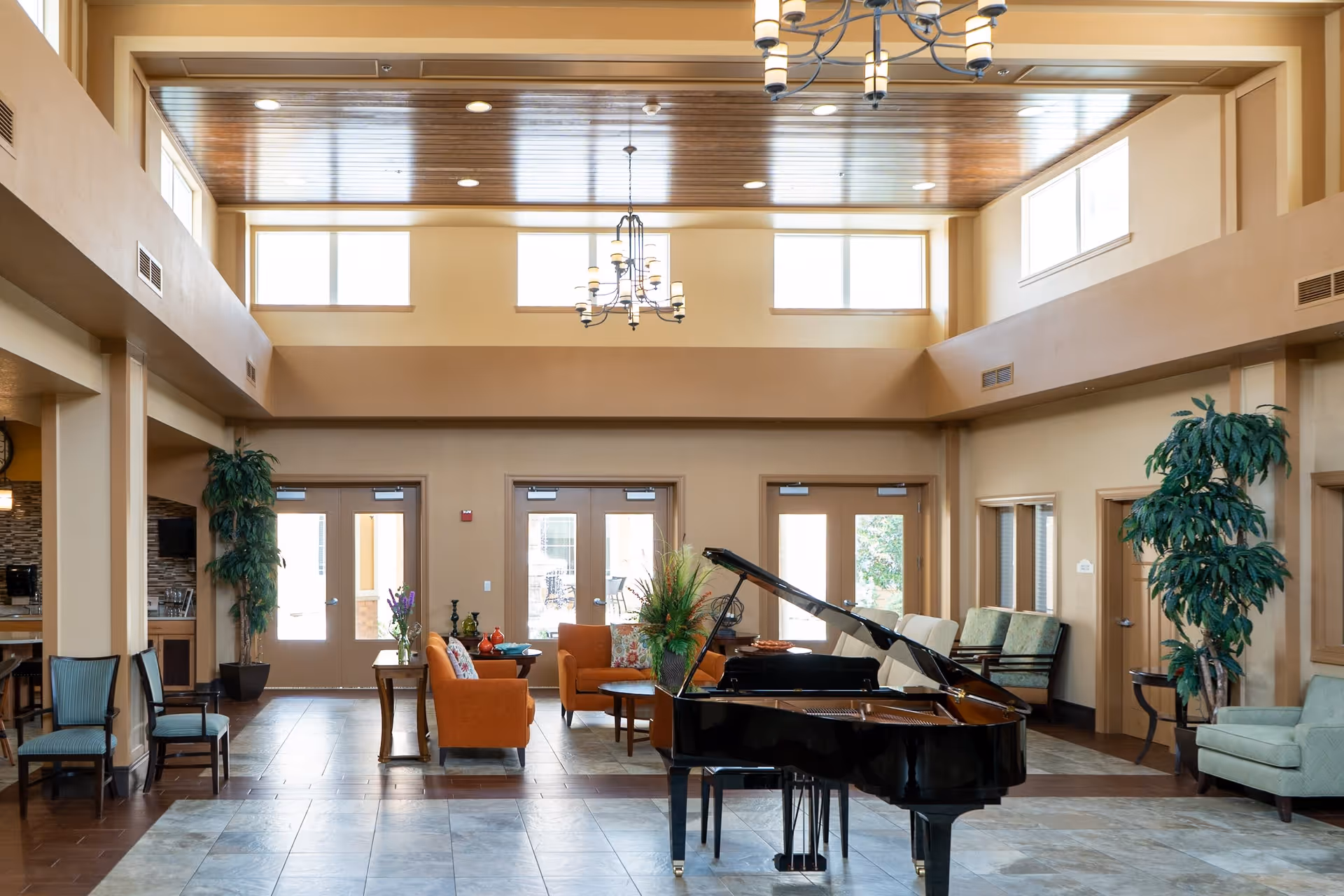 Spacious assisted living facility common area with a black grand piano in the center, surrounded by comfortable seating including orange and green armchairs. The room features high ceilings with wooden paneling, large windows letting in natural light, and decorative plants placed around the space.
