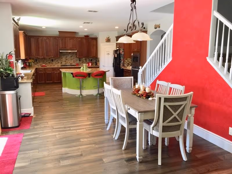 A bright and spacious kitchen and dining area with wooden flooring. The kitchen features dark wood cabinets, a green island with two red bar stools, and modern appliances. The dining area has a wooden table with six chairs, decorated with a centerpiece of candles and autumn leaves. A red accent wall and white staircase railing are visible in the background.