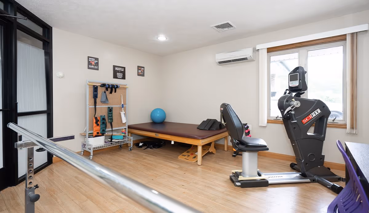 A senior living facility exercise room with a wooden floor, a recumbent exercise bike, a padded therapy table with a blue exercise ball, various exercise bands and equipment on a rack, and a metal handrail in the foreground. The room has a window with vertical blinds and light-colored walls.