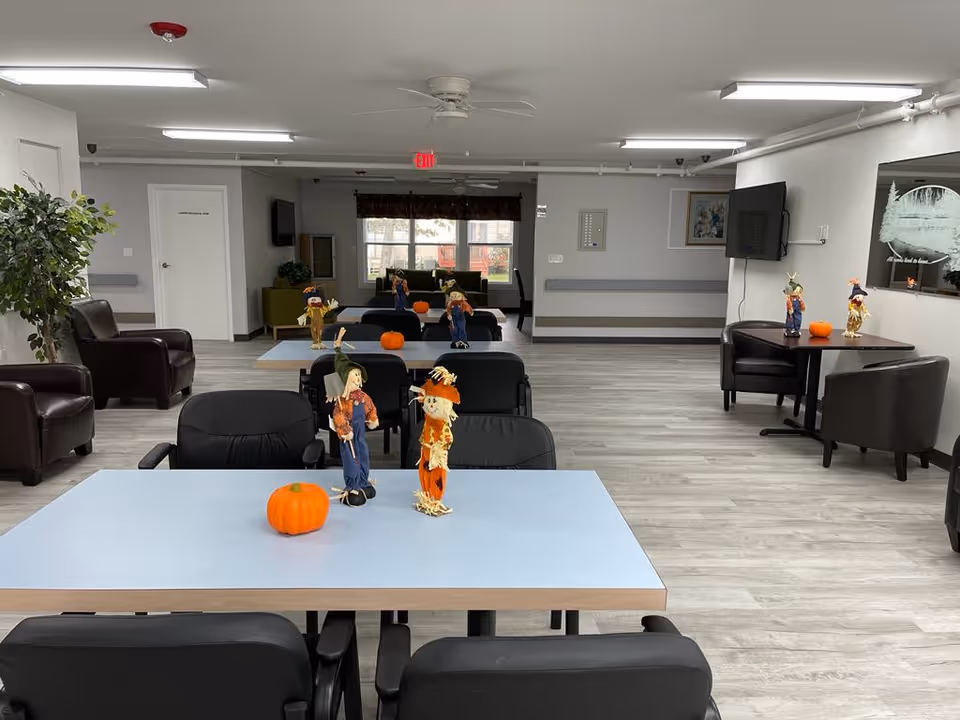 A spacious common area in a senior living facility with multiple tables and black chairs arranged neatly. The tables are decorated with small scarecrow figurines and orange pumpkins, suggesting a fall or Halloween theme. There are several leather armchairs along the walls, a large window at the back letting in natural light, and a wall-mounted TV on the right side. The room has light-colored wood flooring and white walls with some framed artwork.