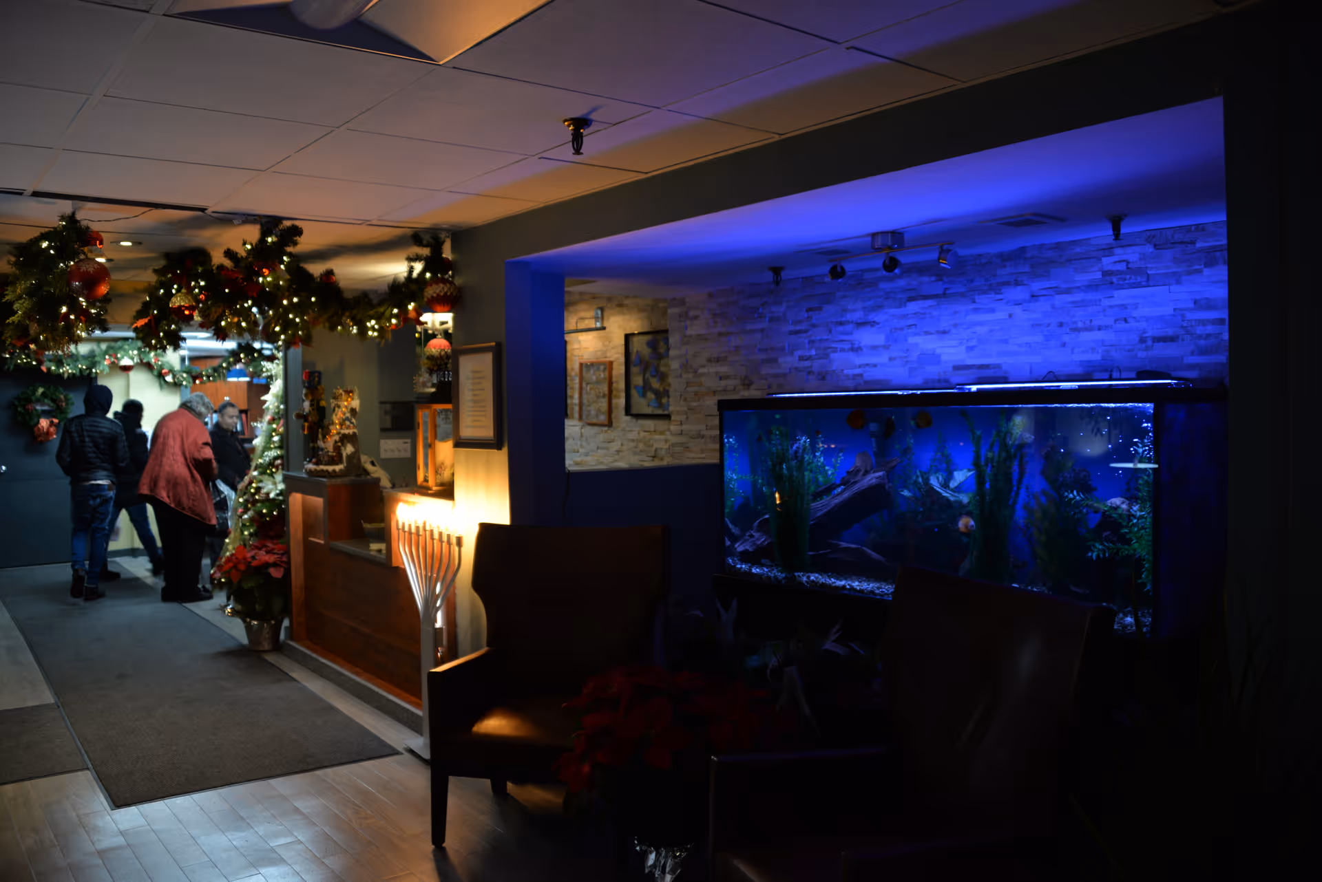 Interior hallway of a senior living facility decorated with Christmas garlands and lights. Several people are walking down the hallway. On the right side, there is a large illuminated fish tank built into the wall with two chairs in front of it. The area is dimly lit with a cozy ambiance.