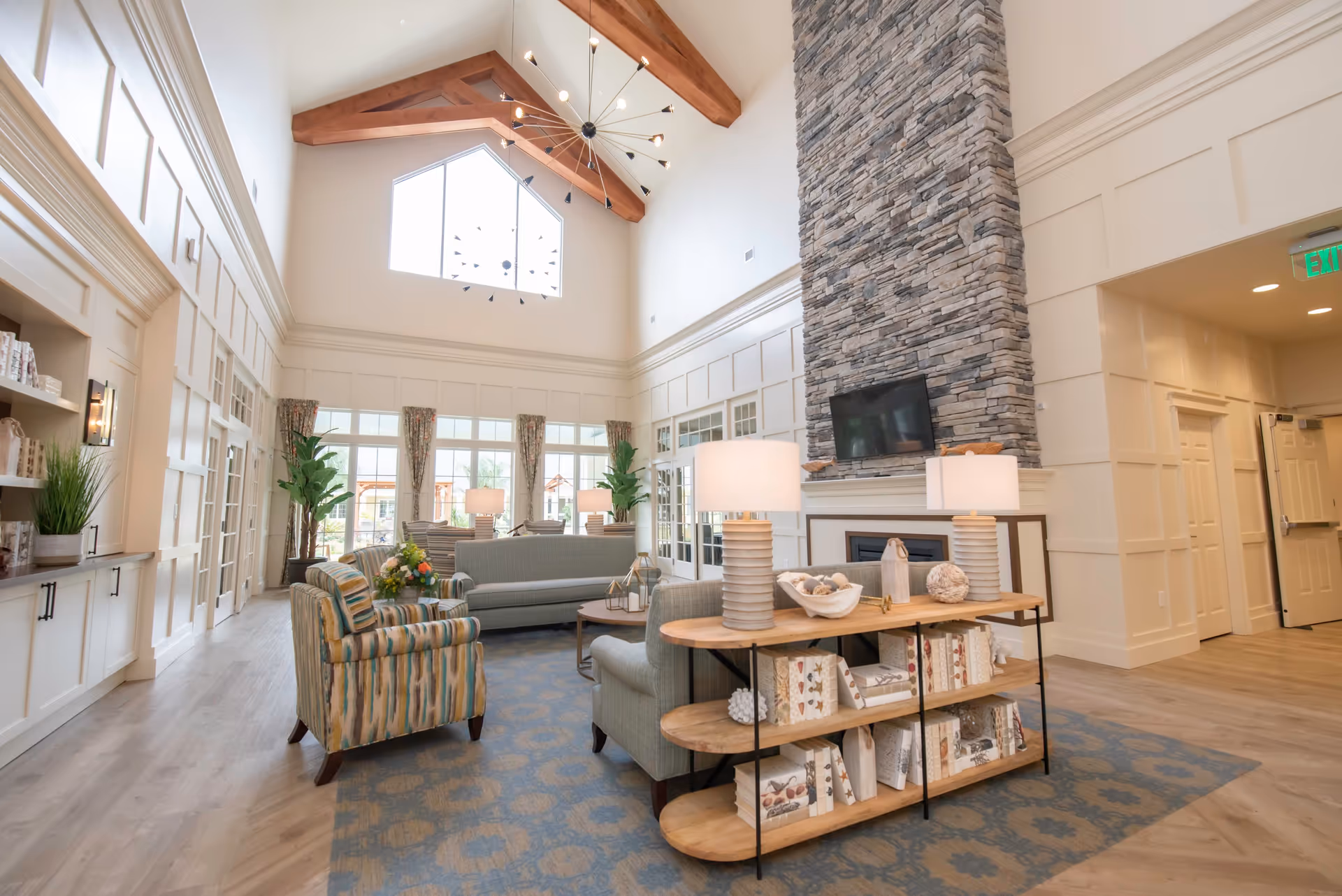 A spacious and bright living room with high vaulted ceilings featuring wooden beams and a large window. The room includes a stone fireplace with a mounted TV above it, multiple sofas and armchairs arranged around a coffee table, a wooden shelf with decorative items and lamps, and large windows with floral curtains letting in natural light. The flooring is light wood with a blue patterned rug.