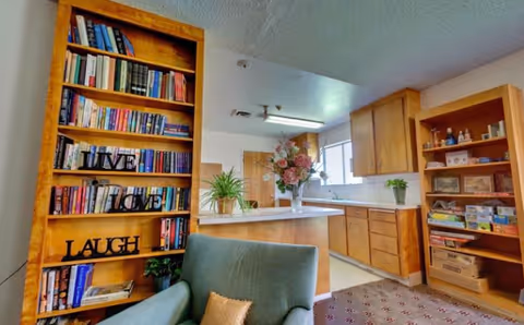 Cozy common room with bookshelves, an armchair, and a view into a small kitchen with wood cabinets.