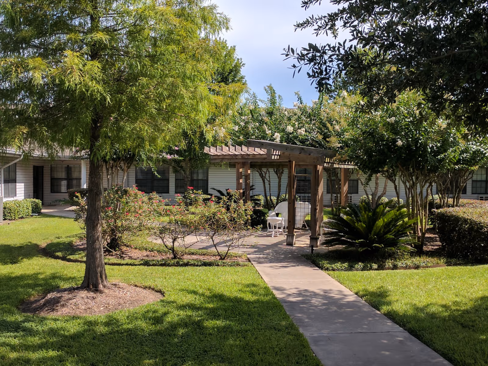 A sunny outdoor garden area at The Courtyards at Pasadena featuring a concrete walkway leading to a wooden pergola with white chairs underneath. The garden is surrounded by green grass, various trees, bushes, and flowering plants. The building with multiple windows is visible in the background.