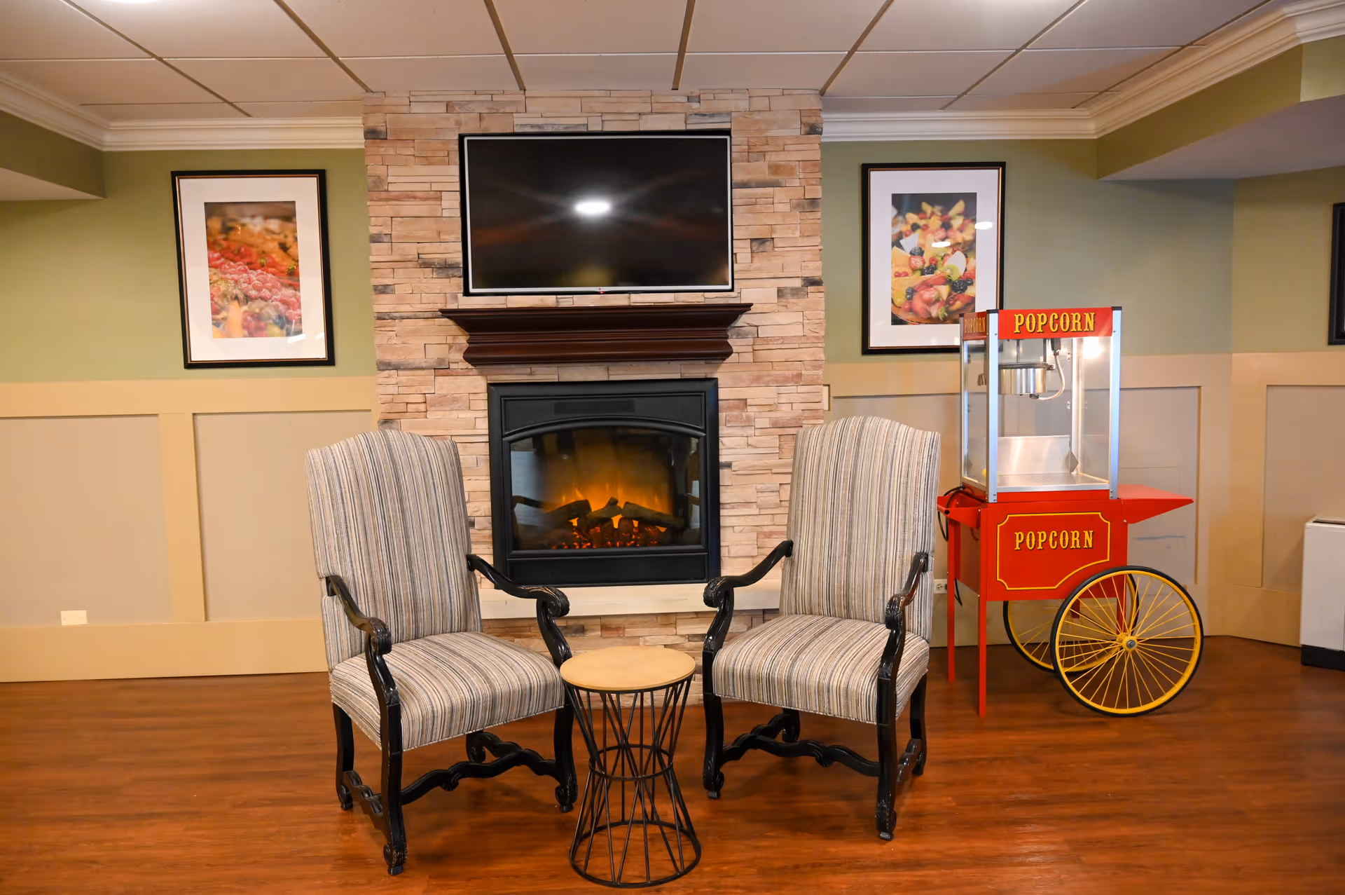 Two striped armchairs with black wooden frames are positioned around a small round table in front of a stone fireplace with a mounted flat-screen TV above it. On the right side, there is a red and yellow popcorn machine on wheels. The walls are painted green and beige with two framed pictures hanging on either side of the fireplace.