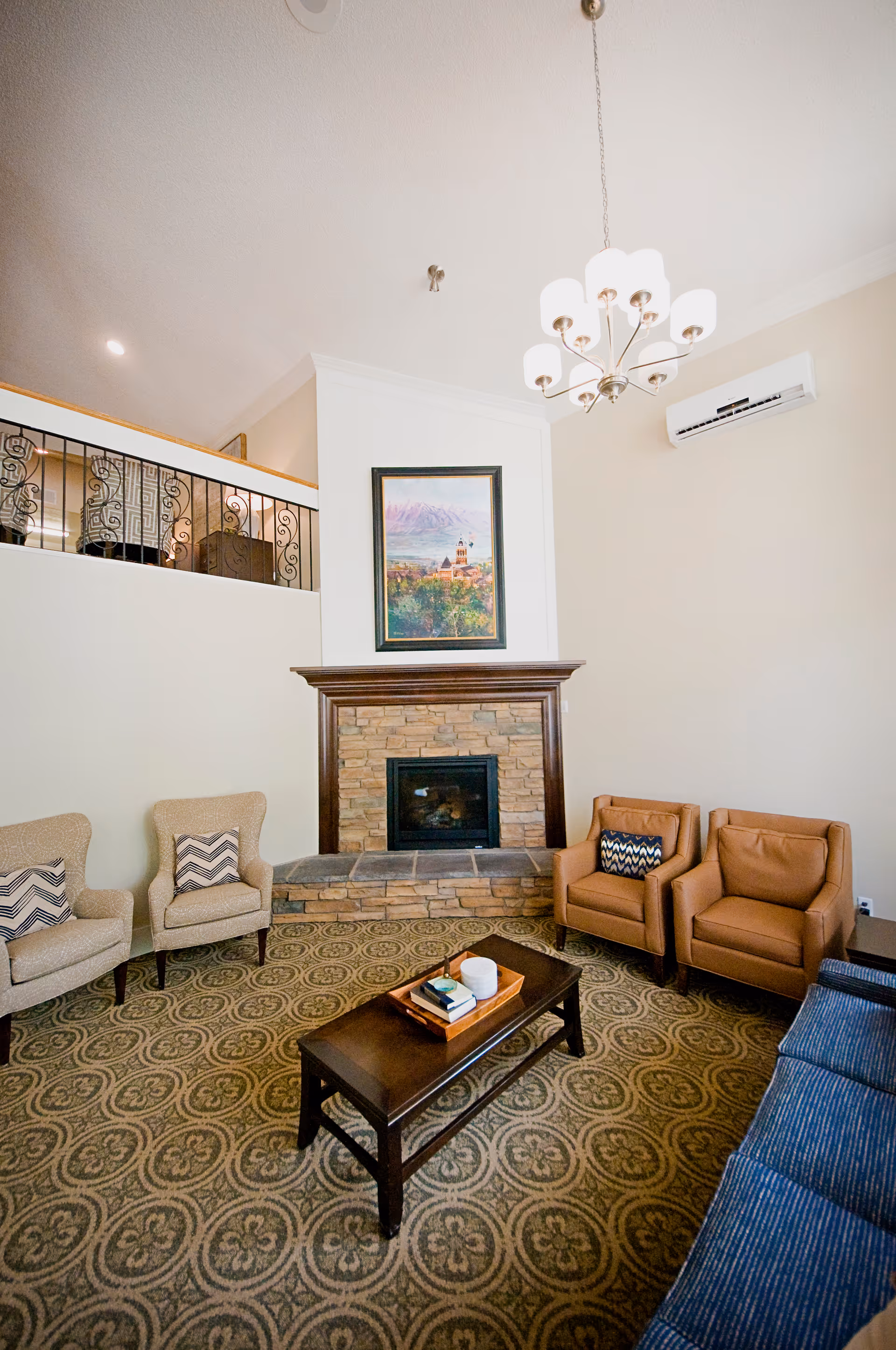 A cozy living room with a stone fireplace centered on the far wall, flanked by two beige armchairs with patterned cushions on the left and two brown leather armchairs with patterned cushions on the right. A dark wooden coffee table with a tray holding books and decorative items sits on a patterned carpet. Above the fireplace hangs a framed painting of a scenic landscape. The room features a high ceiling with a modern chandelier and a wall-mounted air conditioning unit.