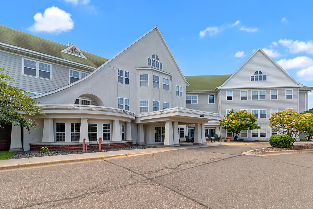 Exterior view of a senior housing facility named Eaglecrest Senior Housing with a large multi-story building featuring multiple windows, a covered entrance, and some landscaping with trees and bushes under a blue sky with scattered clouds.