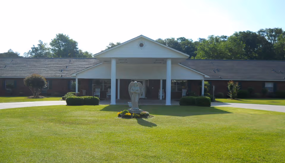 Front entrance of a single-story brick senior living building with a covered portico and an angel statue on the lawn.