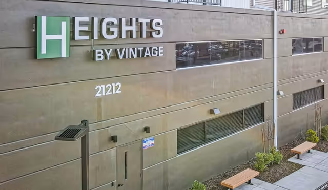 Exterior view of the Heights by Vintage building facade showing a large sign reading 'HEIGHTS BY VINTAGE', the address '21212', windows, and benches along a walkway.