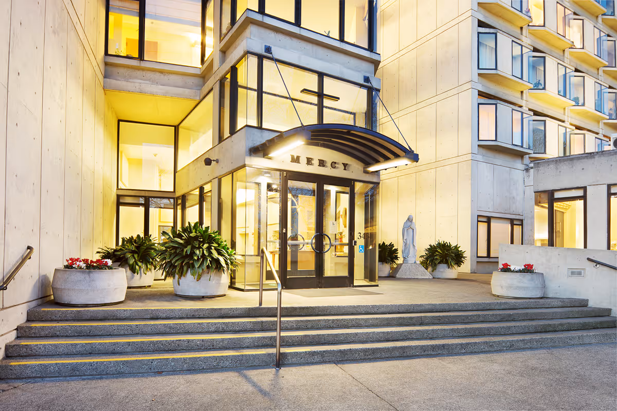 Entrance of Mercy Retirement & Care Center building with glass doors under a curved awning displaying the word 'MERCY'. Concrete steps lead up to the entrance, flanked by large planters with green plants and red flowers. A statue is visible near the entrance, and the building has multiple windows with warm interior lighting.