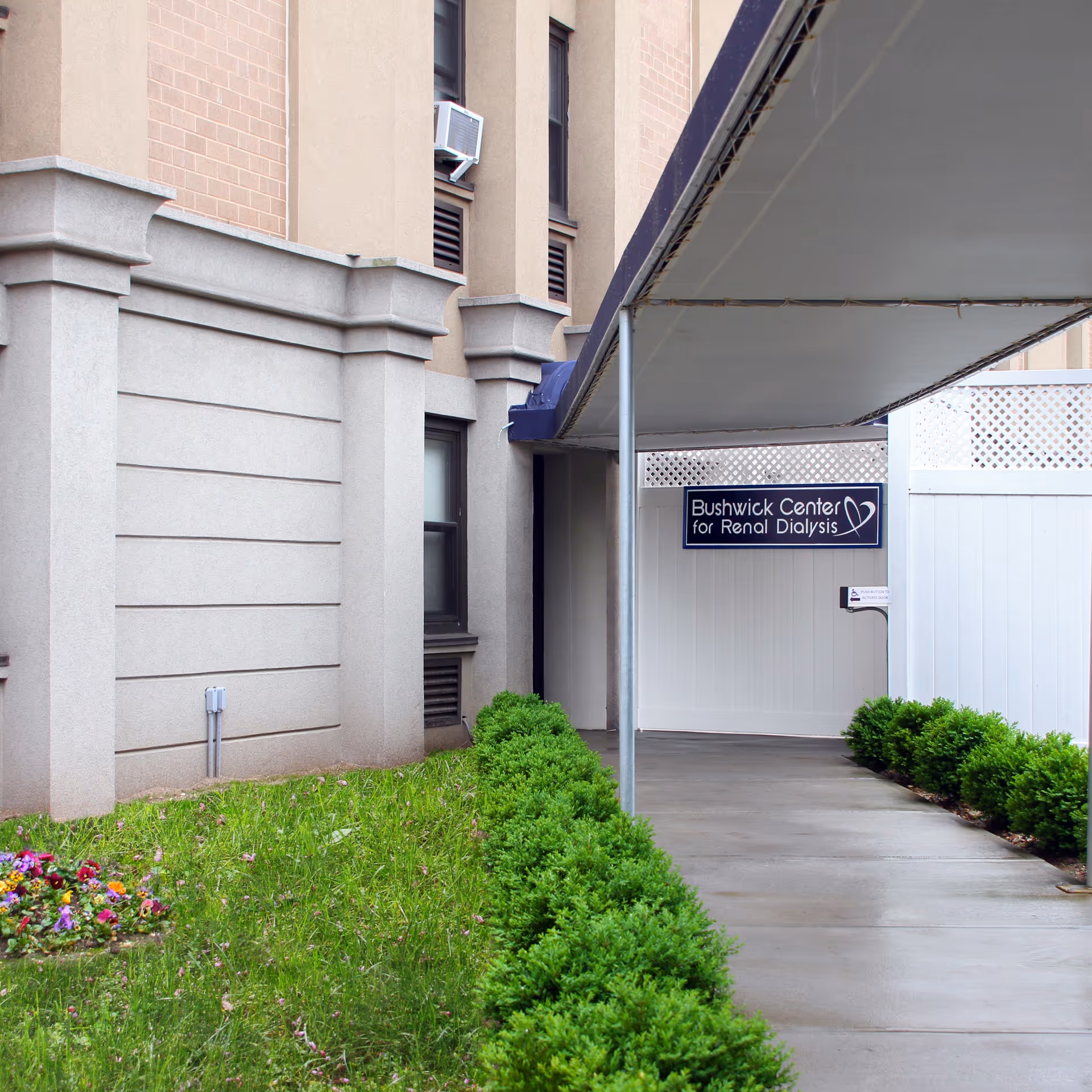 Covered walkway leading to the entrance of Bushwick Center for Renal Dialysis, with green bushes lining the concrete path and a small patch of grass with flowers on the left side. The building exterior is beige with windows and an air conditioning unit visible.